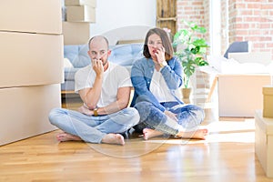 Young couple sitting on the floor arround cardboard boxes moving to a new house looking stressed and nervous with hands on mouth