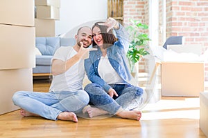 Young couple sitting on the floor arround cardboard boxes moving to a new house smiling making frame with hands and fingers with