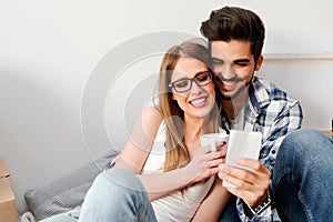 Young couple sitting on the floor of empty apartment looking at smart phone. Move in to new home.