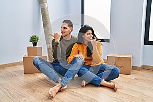 Young couple sitting on the floor at new home smiling with happy face looking and pointing to the side with thumb up