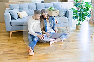 Young couple sitting on the floor of new home using computer laptop and smiling happy for moving to a new home