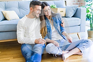Young couple sitting on the floor of new home using computer laptop and smiling happy for moving to a new home