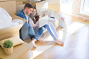 Young couple sitting on the floor of new house around cardboard boxes using laptop, smiling happy for new apartment