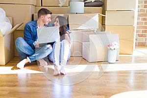 Young couple sitting on the floor of new house around cardboard boxes using laptop, smiling happy for new apartment