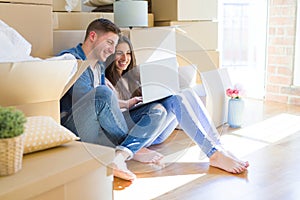 Young couple sitting on the floor of new house around cardboard boxes using laptop, smiling happy for new apartment