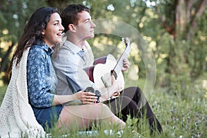 Young couple sitting in the forest and playing guitar, summer nature, bright sunlight, shadows and green leaves, romantic feelings