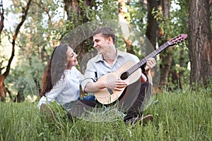 Young couple sitting in the forest and playing guitar, summer nature, bright sunlight, shadows and green leaves, romantic feelings