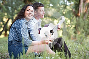 Young couple sitting in the forest and playing guitar, summer nature, bright sunlight, shadows and green leaves, romantic feelings