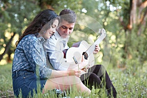 Young couple sitting in the forest and playing guitar, summer nature, bright sunlight, shadows and green leaves, romantic feelings