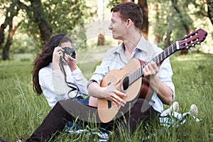 Young couple sitting in the forest and playing guitar, taking photo on old camera, summer nature, bright sunlight, shadows and gre