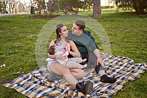 young couple sitting on the grass in the park as they feed their baby and look at each other