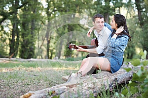 Young couple sitting on a log in the forest and playing guitar, summer nature, romantic feelings