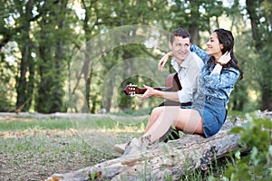 Young couple sitting on a log in the forest and playing guitar, summer nature, romantic feelings