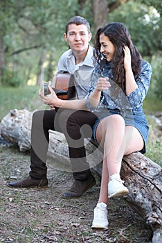 Young couple sitting on a log in the forest and playing guitar, summer nature, romantic feelings