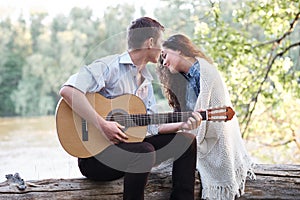 Young couple sitting on a log by the river and playing guitar, summer nature, bright sunlight, shadows and green leaves, romantic