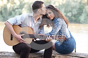 Young couple sitting on a log by the river and playing guitar, summer nature, bright sunlight, shadows and green leaves, romantic
