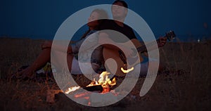 Young couple sitting near bonfire, man playing guitar. Romantic music and happy couple relationships. Romance couple