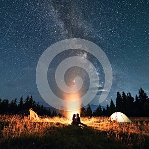 Young couple sitting near campfire under beautiful starry sky.