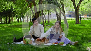 A young couple is sitting in the park on a picnic and stroking their pet dogs on the head. The girl is looking at her