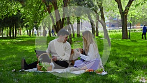 A young couple is sitting in the park on a picnic and stroking their pet dogs on the head. The guy is looking at his