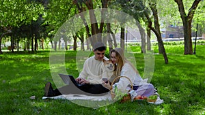 A young couple is sitting in the park on a picnic and stroking their pet dogs on the head. The guy looks at his laptop