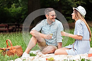 Young couple sitting on a picnic blanket in a park, talking and drinking white wine