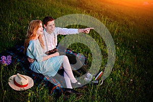 Young couple sitting on the plaid in the park and look at the sunset, the man is pointing to the horizon