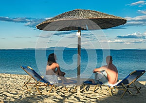 Young couple sitting on a sand and looking to a sea under umbrella. Summer relax on beach.