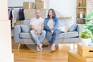 Young couple sitting on the sofa arround cardboard boxes moving to a new house afraid and shocked with surprise expression, fear