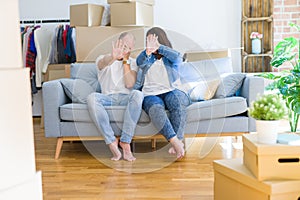 Young couple sitting on the sofa arround cardboard boxes moving to a new house covering eyes with hands and doing stop gesture