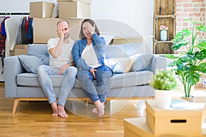 Young couple sitting on the sofa arround cardboard boxes moving to a new house covering one eye with hand, confident smile on face