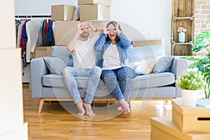 Young couple sitting on the sofa arround cardboard boxes moving to a new house doing ok gesture like binoculars sticking tongue