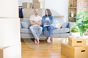 Young couple sitting on the sofa arround cardboard boxes moving to a new house happy face smiling with crossed arms looking at the