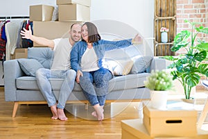 Young couple sitting on the sofa arround cardboard boxes moving to a new house looking at the camera smiling with open arms for