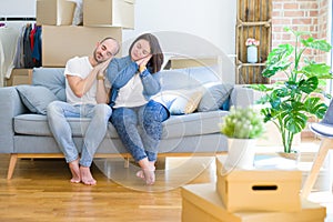 Young couple sitting on the sofa arround cardboard boxes moving to a new house sleeping tired dreaming and posing with hands