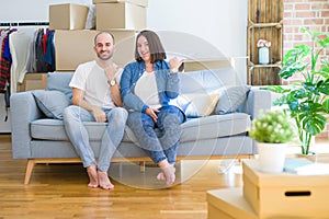 Young couple sitting on the sofa arround cardboard boxes moving to a new house smiling with happy face looking and pointing to the