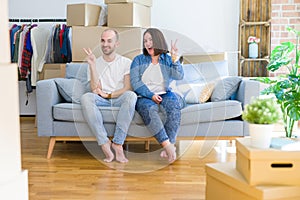 Young couple sitting on the sofa arround cardboard boxes moving to a new house smiling with happy face winking at the camera doing