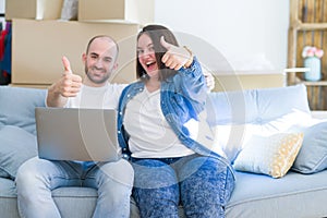 Young couple sitting on the sofa arround cardboard boxes moving to a new house using laptop happy with big smile doing ok sign,