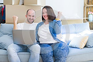 Young couple sitting on the sofa arround cardboard boxes moving to a new house using laptop screaming proud and celebrating