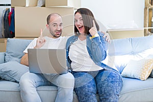 Young couple sitting on the sofa arround cardboard boxes moving to a new house using laptop surprised with an idea or question