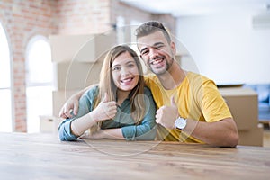 Young couple sitting on the table movinto to new home with carboard boxes behind them doing happy thumbs up gesture with hand