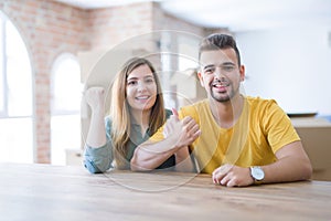 Young couple sitting on the table movinto to new home with carboard boxes behind them smiling with happy face looking and pointing