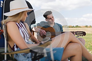 Young couple sitting together in front of van, camping and playing at guitair.