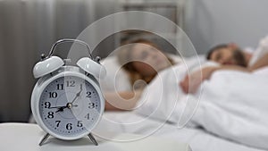 Young couple sleeping in bed with ringing alarm clock on table, sleep biorhythms