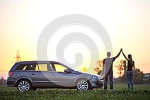 Young couple, slim attractive woman with long ponytail and athletic man holding raised arms at silver car in green field on clear
