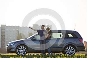 Young couple, slim attractive woman with long ponytail and handsome man pointing in distance standing at silver car in green field