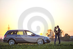 Young couple, slim attractive woman with long ponytail and handsome man standing at silver car in green field on clear sky at