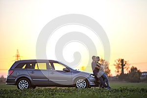 Young couple, slim attractive woman with long ponytail and handsome man standing at silver car in green field on clear sky at