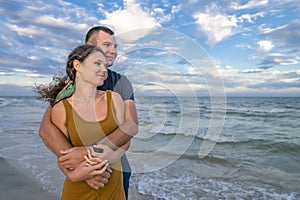 Young couple smile and hug, enjoying vacation at sea. Man and woman relax on evening beach with blue cloudy sky after sunset