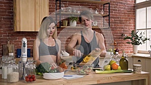 Young couple in sports clothes standing behind kitchen table preparing on lunc fresh salad with vegetables in the